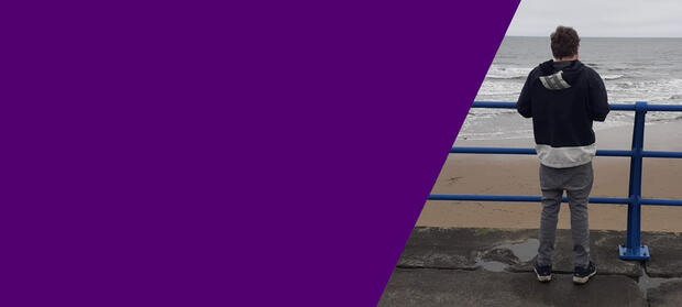 Ben stands with his back to the camera at some railings at a beach, looking out to sea.