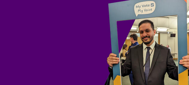 A man wearing a suit holds up a frame with the My Vote My Voice logo