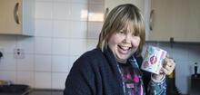 Woman with light hair smiling, stood in kitchen holding a mug near her face.