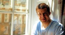 Young man wearing blue top smiling into camera whilst sat next to window.
