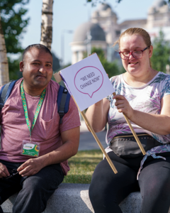 A woman sitting on a wall holding a sign which says 'We need change'. On either side of her are two friends