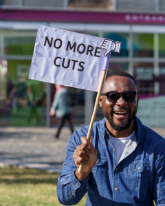 A man in a street holding up a sign which says 'No more cuts'