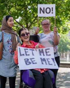 Three woman holding up signs in the street. One sign says Let be be me.