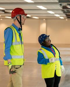A young woman on an apprenticeship is talking to a man on an empty floor of a building. Both are wearing hard hats and high visibility vests