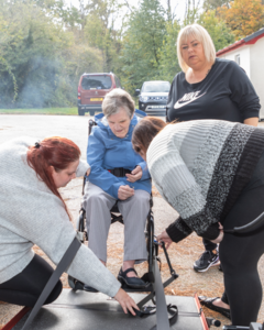 Three support workers are helping a woman in a wheelchair get onto a ramp to get into a mini van
