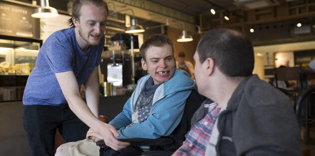 Three men in a pub, one standing reaching out to shake the hand of one of the other two who are seated.