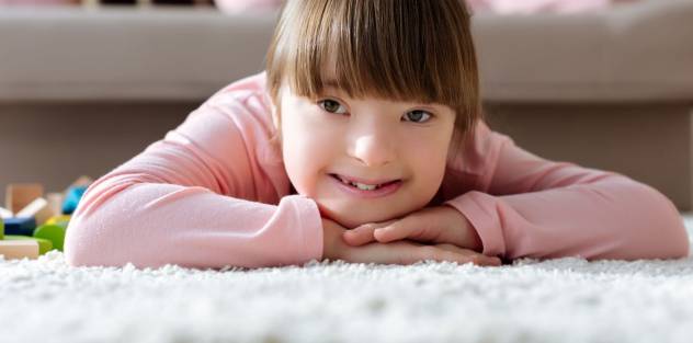 Girl with downs syndrome smiling at the camera lying on a rug with her head on her hands