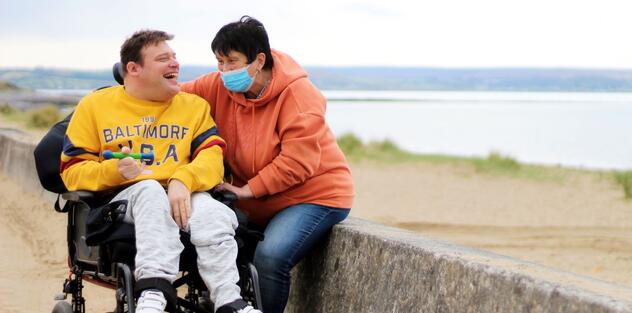A man in a wheelchair is laughing with a woman who is sitting on a wall which overlooks a shingle beach
