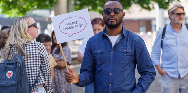 A man holding up a sign saying 'No more cuts' with a crowd of other activists campaigning for change in a park
