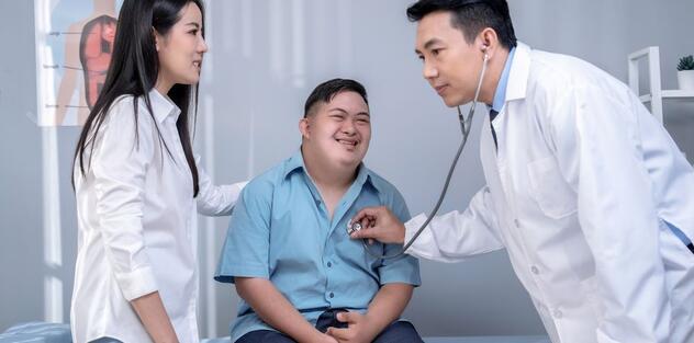 A doctor using a stethoscope on a male patient who has Down's syndrome and is accompanied by his carer